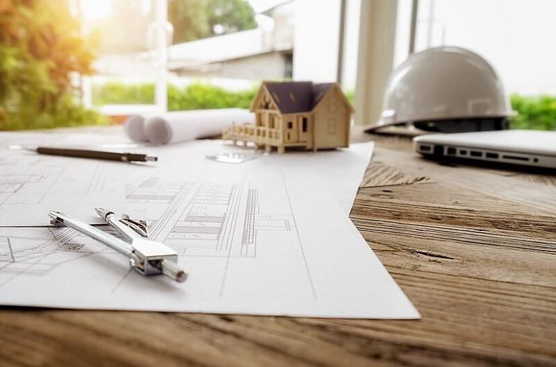 Des plans de construction, un casque de chantier et une petite maison en bois sur une table Le CAUE vous conseille dans votre projet de construction, extension, rénovation